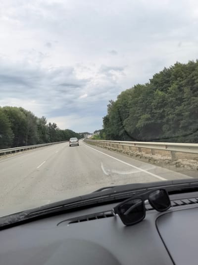 The image captures a view from inside a vehicle traveling on a two-lane road surrounded by dense trees. The sky is overcast with gray clouds. In the distance, a white car is visible driving in the same direction. The foreground shows a portion of the car's dashboard with a pair of black sunglasses resting on it. The road has white lane markings and a metal guardrail runs along the right side. The environment appears to be a rural or semi-rural setting, possibly on a highway. The location is specified as Pigarevskii, Russia.