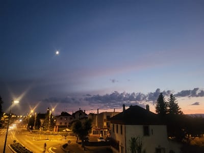 A twilight cityscape is depicted with a dramatic sky transitioning from deep blue to pink hues. In the center, the moon casts a soft glow. Below, a road curves around a roundabout illuminated by streetlights. Buildings of varying heights line the streets, some topped with chimneys. Trees frame the right side of the scene, their silhouettes contrasting against the fading light. One person walks around the roundabout.