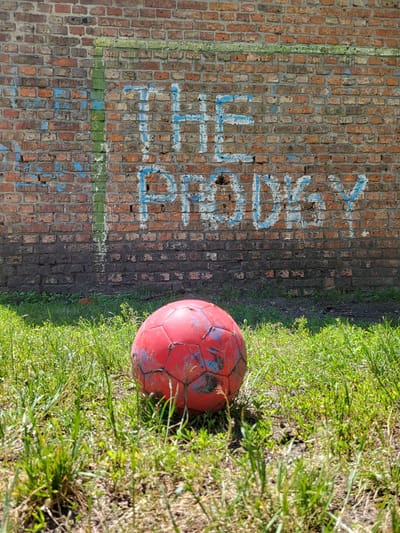 This is a vertical, high-angle shot of a weathered brick wall and a patch of grass. A red soccer ball, showing signs of wear and tear with scuff marks and faded blue accents, rests on the grass to the right of the wall. The brick wall has sections painted with blue and green markings, suggesting it might be part of a sports area. The grass appears somewhat dry and uneven in places, with some patches of bare earth visible.

Visible text in blue spray paint on the brick wall reads "FOOTBALL". There is also a vertical green stripe painted on the left side of the wall and horizontal green and blue stripes towards the bottom.

The lighting suggests it is daytime, with sunlight casting shadows and highlighting the textures of the bricks and grass. The overall impression is of an outdoor, possibly urban or semi-urban, setting where a game of football might be played. The scene feels slightly neglected but still holds the potential for recreation.
