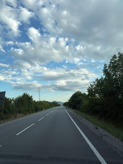 The image depicts a highway in the city of Luhyňa, Slovakia, under a partly cloudy sky. The road is paved and asphalt, with a white line marking the lane. Lush green trees and vegetation line both sides of the road. In the distance, a power line tower is visible. The perspective suggests the photo was taken from a vehicle moving along the highway.