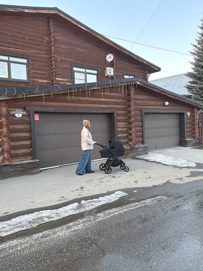 The image depicts an exterior view of Sirenevaya Street, House number 5, in Zelenaya Polyana, Russia. A large, two-story residential structure, constructed from dark brown logs, occupies the left and central portions of the frame. The house features two dark brown roll-up garage doors on the ground level, with multiple rectangular windows visible on both stories. A street sign displaying "СИРЕНЕВАЯ 5" is affixed to the log wall to the left of the left garage door. Decorative icicle-style string lights are strung along the roofline above the garages. An air conditioning condenser unit and a satellite dish are mounted on the upper story facade.
One individual, appearing female, is positioned centrally in the image, walking to the right. She wears a light-colored jacket, wide-leg blue jeans, dark shoes, and a light-colored beanie. She is actively pushing a black, six-wheeled baby stroller with a black canopy.
The ground consists of a concrete or paved sidewalk directly in front of the house, and an asphalt street in the foreground. Patches of melting snow and ice are present on both the sidewalk and the street, with some sections of the asphalt appearing wet. A large evergreen tree is visible on the far right, behind the house, and the sky is clear light blue.