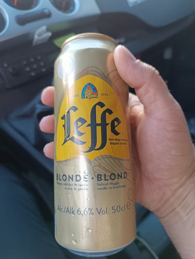 A hand holds a can of Leffe Blonde beer. The can is gold and has condensation on it. The label is yellow and features the Leffe logo, which is a stylized depiction of a church. The text on the label indicates that it is a Belgian Abbey beer with subtle notes of vanilla and cloves. The ABV is 6.6% and the volume is 50cl. The background is blurred and appears to be the interior of a car. The image is taken from a slightly low angle, looking up at the can.