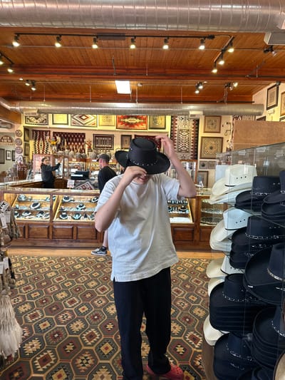 In Oljato-Monument Valley, United States, a young person in a white t-shirt and black pants is trying on a black cowboy hat in a store filled with Native American crafts and hats. The person is standing in the foreground, with their hands on the hat, obscuring their face. To the left, a counter displays jewelry, and behind it, a woman appears to be assisting a customer. Hats are displayed on shelves to the right, with various other items of decor visible throughout the store. The floor is covered with a patterned rug.