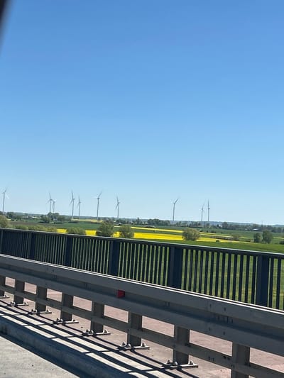 The image shows a view from a bridge in Kończewice, Poland. A blue sky fills the left side of the frame. On the right is a section of the bridge's guardrail, constructed from gray steel and concrete. Beyond the guardrail, a landscape stretches into the distance. A field is striped with green and yellow, indicating different crops or vegetation. Several wind turbines are visible on the horizon, their blades adding a sense of scale to the landscape. The scene is well-lit, suggesting a sunny day.