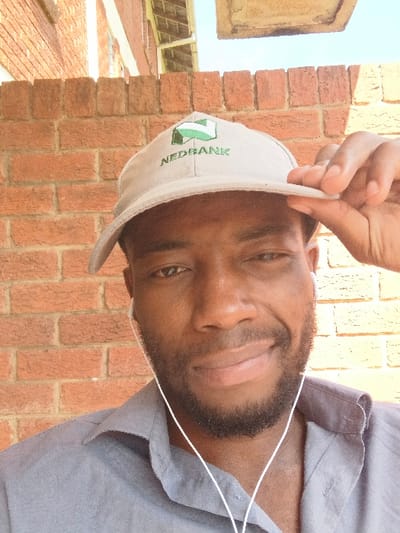 A male individual is depicted in a close-up portrait, directly facing the camera with a slight smile. The subject has dark skin, dark eyes, and a beard. He is wearing a grey collared shirt and a light-colored baseball cap embroidered with a green "NEDBANK" logo. White wired earphones are visible inserted into both ears, with the wires extending downward. His right hand is positioned on the brim of the baseball cap, subtly adjusting it. The background features a red-brick wall, laid in a stretcher bond pattern, occupying the left and upper left portions of the frame. Above the brick wall, elements of a building structure, including a light-colored fascia or guttering, are visible. A segment of clear blue sky is present in the upper right. The scene is illuminated by natural light, casting minor shadows on the subject's left facial area. The image contains no specific architectural landmarks or contextual details to pinpoint a precise location within Chinhoyi, Zimbabwe.