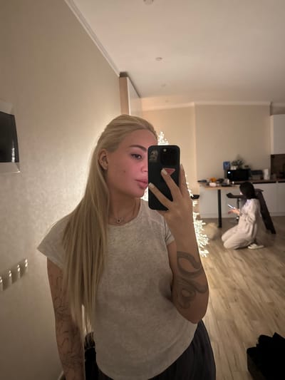 A woman with long blonde hair poses for a selfie, holding a black smartphone. She wears a gray t-shirt, and tattoos are visible on her arm and shoulder. In the background, another person is kneeling near a desk, looking at a device. The room appears to be a modern living space with light-colored walls and wooden floors. A string of white lights is draped across the scene.