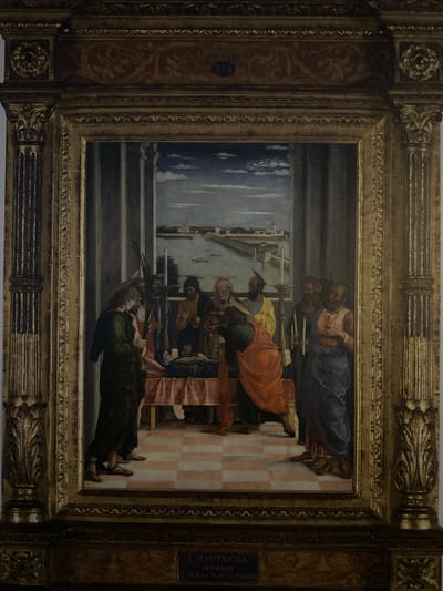 This is a full shot of a framed Renaissance painting depicting a religious scene. The painting is ornately framed with gold accents and detailed carvings. The scene itself shows a group of figures gathered around a figure lying on a bed, possibly a deathbed scene or funeral. The figures are dressed in flowing robes, typical of the period. The background features architectural elements and a distant landscape with a body of water and buildings. The foreground has a checkered floor pattern. At the bottom of the painting, there is a plaque with text, likely an artist's signature and title, which reads "A. MANTEGNA 1431-1506 EL TRÁNSITO DE LA VIRGEN". There is no indication that this painting is located in Puerto De Mazarrón, Spain.