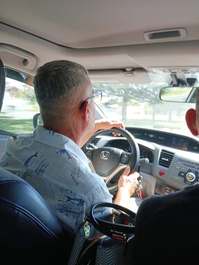 The image shows two people inside a car, with the driver in the foreground. The driver is a man with gray hair, wearing glasses and a light blue shirt with a pattern of dolphins and seaweed. He is holding the steering wheel of a modern car, which appears to be a Honda based on the emblem on the steering wheel. The car's dashboard is visible, including a digital display showing "48" and other information, as well as a radio and air conditioning controls. A red button, possibly for the engine start/stop, is also visible.
The setting is a car interior, likely on a sunny day, given the bright lighting and the appearance of trees and buildings through the windshield and side windows. The time of day appears to be daytime, possibly late morning or afternoon. The overall mood seems casual and everyday, with the focus on the act of driving. A handbag with a checkered pattern and a gold charm is visible on the passenger seat next to the driver. The car is likely in motion, as suggested by the composition and the hands on the steering wheel. The location context provided is Karatay, Turkey, but there are no specific visual cues in the image to confirm this location.