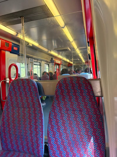 The image depicts the interior of a train carriage, likely a commuter train, featuring rows of seats with a distinctive repeating pattern of interlocking red and blue shapes. The seats are arranged in forward-facing pairs, with a central support dividing them. Passengers can be seen seated in the background, though their features are indistinct. Overhead, fluorescent lighting fixtures are visible. The overall environment suggests a public transportation setting. While the image provides no explicit indication of being in Amadora, Portugal, the seat pattern is consistent with that used by the Fertagus train company in the Lisbon metropolitan area, which includes Amadora. Therefore, the location may be in a train carriage within the Amadora-Lisbon region of Portugal.