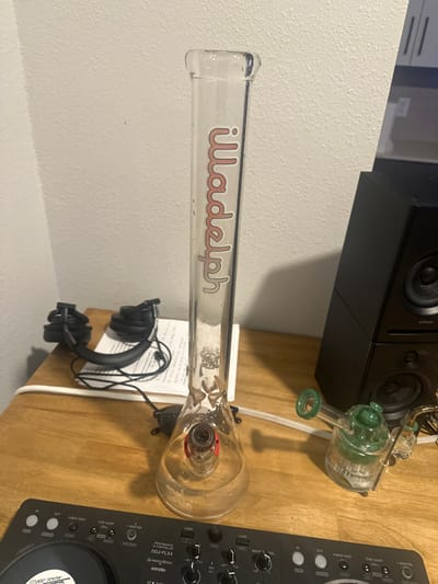 A clear glass beaker-style water pipe, labeled "liladelph" in pink lettering, stands centrally on a light-colored wooden desk in Gainesville, United States of America. It contains water and a reddish-brown bowl with a downstem. To its immediate right rests a smaller, green-tinted glass water pipe or dab rig with intricate percolators. Further right, a black two-tier speaker system is visible. To the left of the main water pipe, a pair of black over-ear headphones with a cable are positioned on a stack of white papers. A white power strip or cable runs horizontally across the desk in front of these items. In the immediate foreground, the top portion of a black Pioneer DDJ-FLX4 DJ controller, featuring a jog wheel with a "SONIC YOUTH" sticker, is visible. The background consists of a plain, light-colored wall. No individuals are present in the scene.