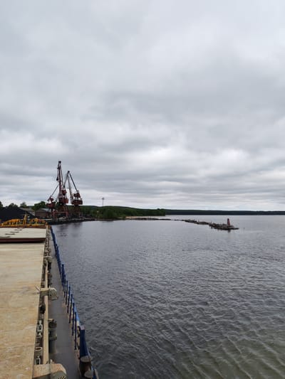 The image captures an industrial port scene on a cloudy day in Medvezhyegorsk, Russia. In the immediate foreground on the left, the weathered and rusty deck of a ship or barge is visible, featuring a blue railing that extends along its side, suggesting the vessel is docked or moving alongside a waterfront.

In the midground to the left, two prominent industrial port cranes, characterized by their rusty reddish-brown and grey steel structures, stand at the dock. Adjacent to the cranes, a large pile of dark material, likely coal, is visible, indicating cargo handling operations. Behind the dock area, a few low buildings are nestled among green trees.

The center and right side of the image are dominated by a wide body of dark water, showing subtle ripples across its surface. Extending from the distant, forested shoreline into the water is a long, low structure composed of logs or wooden pilings. At the far end of this structure, a distinct red and white striped navigational marker or small lighthouse stands, aiding in waterway navigation. The background features a continuous stretch of low, tree-covered land along the horizon, all set beneath a uniform, overcast grey sky with diffuse clouds.

The scene conveys a quiet, working port environment. No individuals are visible, and no specific activity is explicitly taking place. The time of day appears to be daytime, under heavily overcast conditions. No discernible text is present in the image.