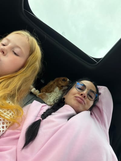The media file shows a selfie taken from inside a vehicle with a sunroof open to an overcast sky. Two people and a dog are visible. In the foreground, a young girl with blonde hair is looking down, her face partially obscured. Behind her, a woman with dark hair, wearing glasses and a pink hoodie, is looking into the camera. Her mouth is slightly open, and she appears to be making a playful expression. A small, light brown dog with long ears is nestled between the woman and the sunroof. The woman has a black braid that rests on her shoulder. The interior of the car appears to be dark. The sky visible through the sunroof is a uniform pale gray, suggesting a cloudy day. No text is clearly visible. The image seems to capture a casual moment of travel. The location is identified as Bronnitsa, Russia.