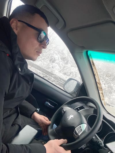 A male individual is observed in the driver's seat of a Peugeot vehicle, positioned in what is identified as Abinsk, Russia. He wears a dark jacket and reflective sunglasses. His right hand is visible on the steering wheel, while a smartphone with a white screen rests on his lap or upper thigh. His gaze is directed forward and to the right, out the windshield. The exterior environment visible through the vehicle's windows displays a snow-covered landscape with heavily coated trees and ground, indicative of winter conditions. The vehicle's interior features a dark dashboard and door panel, with a light gray headliner and sun visor. A turquoise tint is present at the top of the windshield. No other individuals or direct interactions are visible.