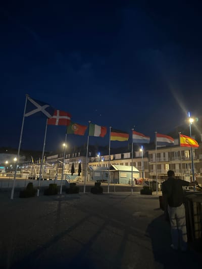 The image captures a view of the waterfront in Saint-Valery-en-Caux, France, at night. A line of flags, including those of Scotland, Denmark, Portugal, Italy, France, Germany, Luxembourg, the Netherlands, and Spain, are flying from poles along the promenade. In the foreground, a person is standing near a dark wooden structure, facing away from the camera and towards the water. Behind the flags, a row of buildings with illuminated windows lines the waterfront. The sky is dark blue with some faint clouds. Streetlights and building lights illuminate the scene.