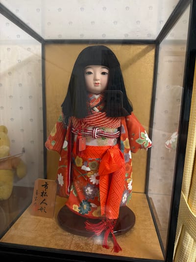 The image shows a Japanese doll displayed in a glass case. The doll is wearing a red kimono with floral patterns and has long, black hair. The doll is standing on a wooden base. There is a small wooden block in front of the doll with Japanese characters on it. The background of the case is a gold color. The image was taken indoors, likely in a home or museum. The location is Kawasaki, Japan.