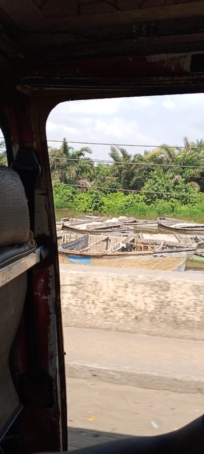 The image depicts a static scene viewed from inside a stationary or moving vehicle, evidenced by the visible door frame, seat, and interior roofline on the left and top portions of the frame. The exterior scene, located in Ketu Adieowe, Nigeria, features a body of water in the midground, likely a river or canal, populated by approximately ten wooden boats. These boats are small, simple vessels, some appearing to be partially submerged or in various states of disrepair. One prominent boat in the central foreground has a tan hull with a distinct horizontal blue stripe and a smaller red detail.

Beyond the water, a dense line of tropical vegetation, including numerous palm trees and lush green bushes, forms the background. Several horizontal power lines or communication cables are visible traversing the sky above the vegetation. The sky is partially cloudy with white cumulus formations against a lighter blue expanse. The immediate foreground outside the vehicle consists of a light-colored, possibly sandy or unpaved, ground surface. No individuals are visible in the exterior scene; the only indication of human presence is the interior of the vehicle from which the photograph was taken.