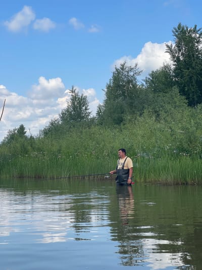 A man is standing in shallow water, wearing waders and holding a fishing rod. He is facing to the left, with a slight smile on his face. The water is calm, with reflections of the sky and surrounding vegetation. The background is filled with lush green reeds and trees, with a blue sky and white clouds above. The scene appears to be a natural, rural setting, possibly in the vicinity of Nezhinka, Russia, a village known for its natural beauty and agricultural landscapes. The man is engaged in fishing, a common recreational activity in such environments. The overall atmosphere is peaceful and serene, suggesting a pleasant day spent outdoors.