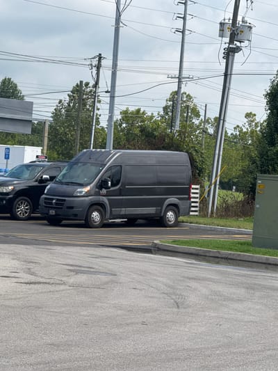 The image depicts an outdoor scene on an overcast day. In the foreground, a gray asphalt parking lot is visible, marked with yellow lines. To the right, a tall, green electrical transformer box stands. Behind the transformer box, there is a patch of green grass and dense foliage. Dominating the mid-ground is a dark gray cargo van parked in a parking space. To the left of the van, a dark-colored SUV is also parked. In the background, several utility poles with numerous electrical wires stretch across the frame. Trees with green foliage line the horizon. A white building with a blue sign that says "EV" is partially visible to the left of the SUV. The scene appears to be a parking area, possibly at a commercial or public facility, in Knoxville, United States of America.