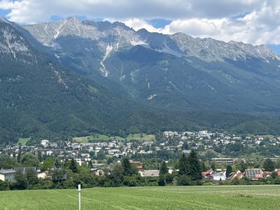 This is an eye-level, medium shot of a wide landscape with a city and mountains. In the foreground, a mown green field is visible with a white marker post. In the mid-ground, a densely populated residential area is nestled at the foot of a mountain. The buildings are a mix of styles and sizes, with some featuring solar panels on their roofs. Tall green trees are interspersed among the houses. In the background, a massive mountain range dominates the skyline, with rugged, rocky peaks partially covered in snow and scree slopes. The sky is partly cloudy with patches of blue. The scene appears to be taken from a vantage point overlooking the city of Innsbruck, Austria, likely from a hillside or elevated area. There are no people or animals visible in the frame.