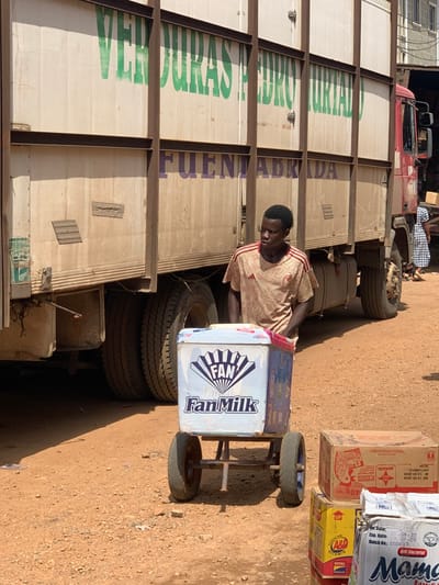 A young man wearing a light-colored shirt with a pattern stands in front of a large truck with a white cab and a tan cargo box. The man is pushing a blue and white cart with the words "Fan Milk" on the front. The cart is parked next to a stack of cardboard boxes with brown and red writing on them. The truck has a sign on the side that reads "Verduras Pedro Turtado" and "Fuente Abrida". The man is pushing the cart on a dirt road, presumably in the town of Ondo, Nigeria, which is located at the coordinates 7.251, 5.196. The image appears to be taken in a commercial area. The man appears to be selling the "Fan Milk." The truck appears to be a delivery vehicle. The scene is brightly lit and the day is sunny.