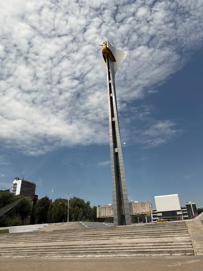 A large, metallic monument topped with a golden statue of a winged figure stands tall against a cloudy blue sky. In the foreground, wide concrete stairs ascend towards the monument. Behind the stairs, several buildings of varying architectural styles are visible, including a tall white building with scaffolding, trees, and a modern building with a large, white facade. A yellow bus is seen on the street in front of the modern building. The scene appears to be in an urban setting, possibly a public square or park.