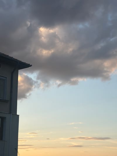 The image shows a building facade on the left, with a grey downspout running vertically along its side. The building is light-colored and has at least one window visible. The roofline extends from the top left of the frame. The majority of the image is occupied by the sky, which displays a mix of clouds. The upper portion of the sky is filled with large, dark grey clouds interspersed with areas of lighter grey and hints of yellow and orange, suggesting the presence of sunlight breaking through. Toward the horizon, the sky transitions to a lighter blue, with scattered, thin, horizontal clouds tinged with yellow and orange, indicative of sunset or sunrise. There are no people or visible actions depicted in the image. The scene is likely set in Belogradchik, Bulgaria, as specified, with the building and sky being the only discernible elements.