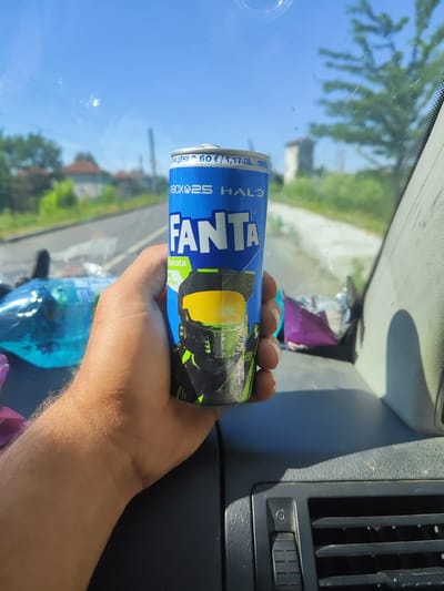 A close-up, first-person perspective shot shows a hand holding a blue and green Fanta can featuring the Xbox logo and Halo imagery. The can's text indicates a price of "60 € / 1,17 лв.". In the background, blurred out, is a street scene with houses and trees under a clear blue sky, suggesting an outdoor, possibly rural, location. The can is positioned in the foreground, obscuring part of the dashboard of a vehicle, which includes a dark gray vent and a portion of a gray pillar. The lighting suggests it is daytime. The scene is likely captured from inside a vehicle traveling through the city of Roza, Bulgaria, on a sunny day.