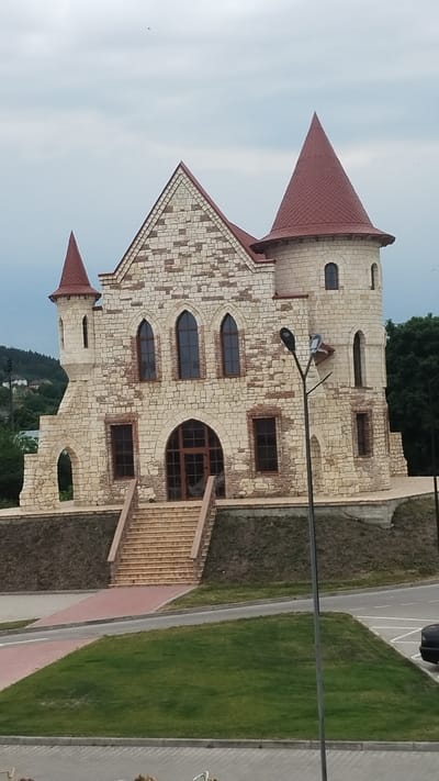 The image depicts a fairytale-like stone building resembling a castle, situated in Berezhany, Ukraine. The castle features a prominent red conical roof atop a tower and a similarly colored pitched roof over the main structure. The exterior walls are constructed from rough-hewn, light-colored stones, interspersed with some darker stones. Numerous arched windows, some with a rounded top and others with a pointed Gothic arch, are visible, many of which are dark-tinted. A large, arched wooden door is present on the right side of the building, accessible via a set of brick stairs. Another set of stairs leads up to the main entrance. To the right of the castle, a well-maintained grassy lawn runs alongside a paved pathway and a road. A black car is partially visible on the road. The sky is overcast and pale blue, suggesting it is daytime with possibly diffused sunlight. The overall scene evokes a sense of fantasy and perhaps a tourist attraction or a unique architectural structure. No people are clearly visible in the frame, and no text is discernible.