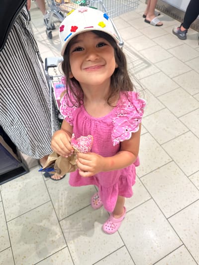 The image shows a young girl in a bright pink dress and a white hat, holding a pink donut with sprinkles. She is smiling at the camera. The girl is in what appears to be a grocery store, with a shopping cart visible in the background. The floor is tiled with a pattern of light-colored squares and speckled flecks. The image is taken from a slightly high angle, looking down at the girl. The lighting suggests it is daytime, and the setting appears to be indoors. The girl's expression is happy and playful. There is no visible text in the image.