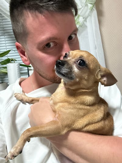 A man is lying down, facing the camera, holding a tan Chihuahua dog in his arms. The dog is looking away from the camera. The man has short, dark hair, light skin, and stubble. Both are wearing white clothing. A houseplant and a white wall are visible in the background. The location is not determinable from the image.