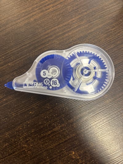 This media file contains an overhead shot of a correction tape dispenser on a dark, wood-grained surface. The dispenser is clear plastic with blue internal components, including visible gears. Text on the dispenser reads "5mm x 16m" and "TORUB TWINGO". The wood grain has a warm, brown tone and appears to be a tabletop or desk. The lighting suggests an indoor setting, with no indication of time of day or weather. There are no people or animals present. The overall scene is a simple, uncluttered depiction of a common office or school supply. The location context of Līvāni, Latvia, does not offer any visual cues within the image itself.
