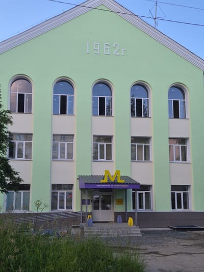 This is a vertical, eye-level shot of a light green, multi-story building in Petrozavodsk, Russia. The building has arched windows on the upper floors and rectangular windows on the lower floors. The year "1962" is embossed on the left side of the building. A purple awning with yellow letters spelling out "MV" is visible above the entrance on the right side of the building. The text below the awning reads "пространство от росмолодежи". The scene appears to be set in the daytime, under a clear sky. There are no people visible in the image.