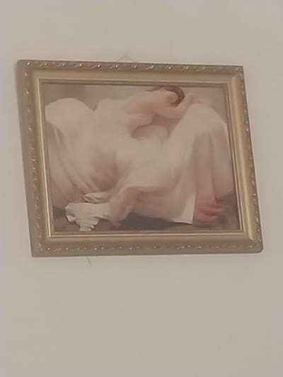 The image shows a framed painting with a gold-colored ornate frame hanging on a white wall. The painting depicts two figures, seemingly human, draped in flowing white and pink fabric. Their bodies are intertwined, with one figure's head resting on the other's shoulder or chest. The style of the painting appears to be classical or romantic. There are no discernible elements in the image that can be used to identify its specific location in Akhalkalaki, Georgia, or any urban environmental context.