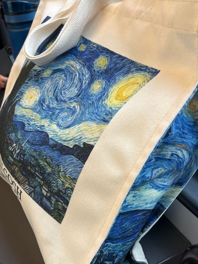 The image shows a tote bag featuring Vincent van Gogh's "The Starry Night" painting. The bag is made of light-colored canvas with the painting printed on the front. The painting depicts a swirling night sky with a crescent moon, stars, and a village landscape below. The handles of the bag are visible on the left side. The background is blurred, revealing parts of what appear to be car or airplane seats. It is not possible to determine the location of the bag in Indjija Municipality, Serbia, based on the visual information provided.