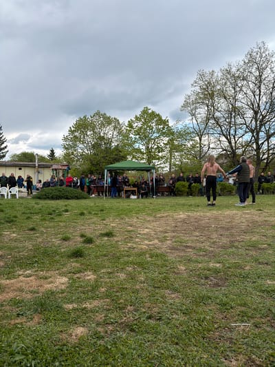 The image appears to be taken in a park-like setting, possibly in or near Dulevo, Bulgaria. The sky is overcast. In the distance, a single-story building is visible. Groups of people are gathered. One group is seated under a green canopy. Two men are standing on a grassy field with their torsos exposed; a third man, wearing a padded vest, is also present.