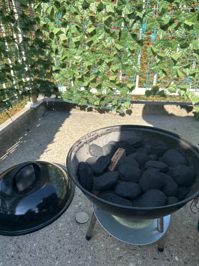 A charcoal grill filled with briquettes sits on a concrete patio in Katwijk, Netherlands. A black lid rests on the ground next to the grill. The grill is surrounded by a decorative green vine-covered fence. The sun casts shadows on the patio, indicating it is daytime.