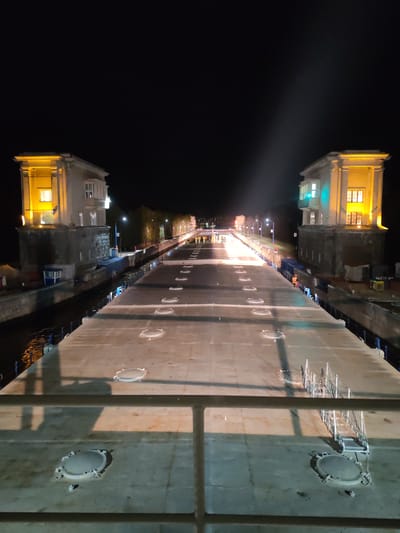 Captured at night, the image presents a straight elevated concrete structure flanked by waterway locks in Водник Снт, Russia. Two illuminated, multi-story control towers bookend the structure on either side. The concrete surface features several circular access points and a metal ladder. Further down the structure, a group of people wearing bright attire can be seen clustered near lighting fixtures. The environment is dimly lit, with street lights and distant buildings visible.