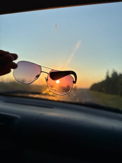 The image shows a pair of sunglasses held up against a sunset. The sunglasses are partially obscuring the sun, which appears as a bright, glowing orb. The lenses of the sunglasses are tinted pink and purple, reflecting the warm hues of the sunset. The background shows a blurred landscape of trees and a road, suggesting the photo was taken from a moving vehicle. The sky transitions from a pale blue at the top to a vibrant orange and yellow near the horizon. A hand is visible at the bottom of the frame, holding the sunglasses. The overall mood is serene and contemplative, capturing a moment of natural beauty. The time of day is clearly sunset, indicated by the low angle of the sun and the warm, diffused light. The location cues, such as the trees and road, suggest a rural or suburban setting. There is no visible text in the image.