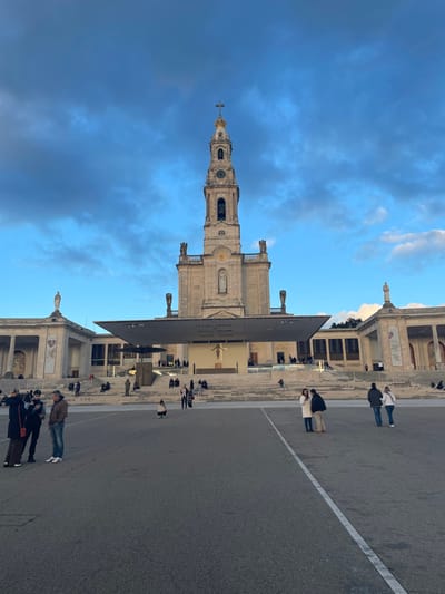 The image displays the Sanctuary of Fátima in Fátima, Portugal, under a partly cloudy blue sky. The central subject is the Basilica of Our Lady of the Rosary of Fátima, a light-colored stone building with a prominent, ornate bell tower topped by a cross. The tower features a clock face and visible bells. Below the tower, the basilica's facade includes statuary and architectural details. In front of the basilica, a modern, dark-colored canopy structure extends over a wide set of steps, beneath which a large crucifix is visible. To the left and right of the basilica are other colonnaded stone buildings, part of the larger sanctuary complex, with a statue visible on the roofline of the structure on the left.
The foreground is a vast, dark grey paved esplanade with faint white ground markings. Scattered across this area and on the steps are numerous individuals and small groups of people. In the foreground-left, a group of four individuals stands together, three appearing to be engaged in conversation. One person wears a dark coat, another a brown jacket and blue jeans. In the midground-right, a couple stands, one person in a light-colored coat, the other in a dark jacket. Further right, another couple walks away from the camera. A single individual is crouched or kneeling on the paved ground in the center midground. Many more people are visible sitting and standing on the steps leading up to the basilica and the modern canopy structure. The overall scene suggests moderate foot traffic within a large, open religious complex.
