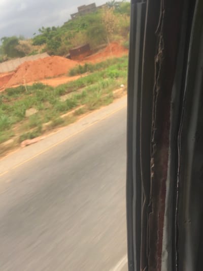 A blurred landscape is seen from a moving vehicle. The top of the frame shows the vehicle's interior. Outside, a road occupies the right side, its gray surface suggesting motion. To the left, a steep bank is visible, with patches of red earth and green vegetation. In the distance, a building sits atop the terrain, partially obscured by vegetation. The blurry quality of the image indicates speed.