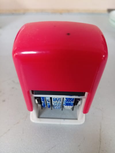 A red date stamp is placed on a light-colored, speckled surface. The stamp has a white base and a black ink pad visible at the bottom, with blue numbers and letters on rotating wheels. The background is out of focus, showing a light-colored wall and possibly part of a table or counter. The image does not provide any information about the city of Rezina, Moldova, or any other location.