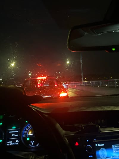 The image shows a night-time car interior view. A rearview mirror is visible on the left, reflecting lights outside. The dashboard features a media display with Bluetooth connectivity, and a speedometer showing 23 km/h. A steering wheel is in the lower right. Outside temperature is 8 degrees Celsius, and the odometer reads 283,823 km. Red tail lights are prominent in the windshield view, indicating traffic ahead. The windshield is wet, reflecting exterior lights.