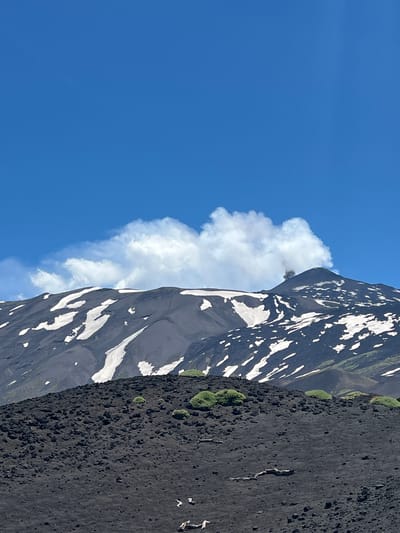 This image depicts Mount Etna, an active volcano located near Linguaglossa, Italy. The foreground is dominated by a stark, black volcanic landscape, likely composed of solidified lava and ash, with sparse patches of green vegetation. In the mid-ground, the slopes of Mount Etna rise, characterized by dark, barren rock interspersed with significant patches of white snow. A plume of dark gray smoke or ash is visible emerging from the summit of the volcano, indicating ongoing volcanic activity. The background consists of a clear, vibrant blue sky with a few scattered white clouds. The overall scene conveys a sense of natural power and the dramatic interplay between volcanic forces and the elements. There are no people visible in the image.