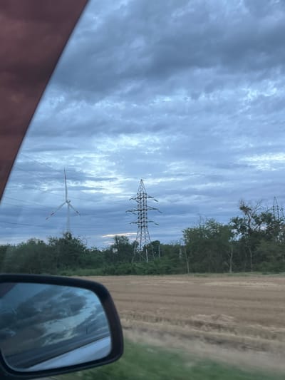The image depicts an outdoor scene, likely taken from a vehicle, showing a power line tower and a wind turbine against a cloudy sky. The sky is filled with dramatic, streaky clouds in shades of gray and white, suggesting a dynamic weather system. In the background, there's a line of trees bordering a field of dry, light-brown earth, possibly a harvested agricultural field. The scene is captured from a low angle, with the top of a power line tower visible in the upper right quadrant and another, larger power line tower in the center. A wind turbine with white blades and red tips is positioned in the lower center of the frame, its blades appearing to be in motion. A side-view mirror of a vehicle is in the lower right corner, reflecting the sky and clouds. The overall impression is one of a rural or semi-rural landscape under a moody, overcast sky, possibly during twilight. The image does not contain any visible text.