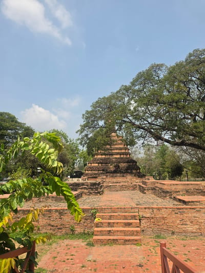 The image shows the ruins of Wat Yai Chaimongkol in the Historic City of Ayutthaya, Thailand. The main subject is a large brick stupa, partially surrounded by trees and greenery. In the foreground, lush green foliage is visible. The sky is light blue with scattered clouds. A portion of a red-orange metal structure is visible on the right side of the image. The image is taken from a low angle, looking up at the stupa.