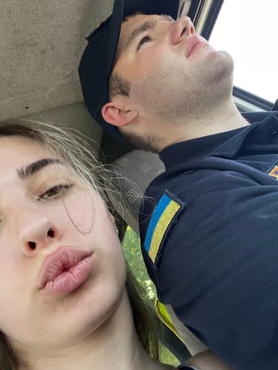 A close-up, horizontal, selfie-style photo shows two people, a man and a woman, lying down together. They are positioned so their heads are in the foreground and their bodies extend away from the camera. The man is on the left, wearing a dark blue uniform with a Ukrainian flag patch on the sleeve. He is wearing a black baseball cap and has a stubble beard. His eyes are closed, and his head is tilted upwards. The woman is on the right, with her head in the foreground. Her lips are slightly parted as if she is about to kiss the camera. Her eyes are open, and she is looking towards the camera. Her hair is light brown and falls around her face. The background appears to be a car interior, with a gray fabric seat visible in the bottom left and a hint of green foliage through a window on the right, suggesting an outdoor setting. The lighting suggests it is daytime. The overall mood seems intimate and relaxed.