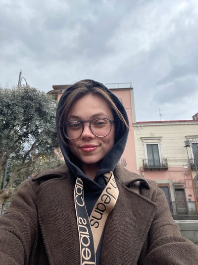The image depicts a female individual in a medium close-up, selfie-style shot, set outdoors in the city of Scafati, Italy. The subject is a young adult female with light skin, dark hair partially covered by a dark hooded sweatshirt, and is wearing round, brown-framed glasses. She is smiling subtly, displaying dimples on both cheeks. Her attire includes a dark brown, possibly herringbone-patterned, collared coat over the dark hooded sweatshirt. A beige and black strap or lanyard, printed with "Calvin Klein Jeans" text, hangs around her neck, visible over her coat.

The background on the left features a dense-leafed tree, possibly an olive tree, with a dark streetlamp or utility pole partially visible behind it. On the right, two multi-story buildings are present under an overcast sky. The prominent building has a light pink facade with a visible railing on its flat roof. Adjacent to it on the right is a lighter-colored building, cream or light beige, featuring ornate white architectural trim, a window with a small iron-railed balcony, and two brown wooden doors below. A television antenna is visible on the roofline of this lighter building. The sky is uniformly grey and cloudy.