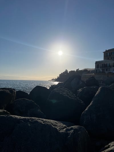 The image shows a view of the sea and sky taken from a rocky breakwater in Bogliasco, Italy. The sun is bright in the sky on the left, casting long rays of light. Buildings are visible along the coast, slightly faded due to the bright light. The breakwater is composed of large, dark rocks. The sea appears calm. The photo is taken with the camera rotated.