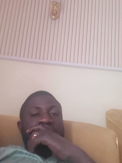 An adult male with dark skin, short dark hair, and a beard is depicted in an indoor environment in Babawa, Nigeria. He wears a light green shirt and a ring with a red stone on his right ring finger. His right hand is positioned with fingers partially covering his mouth and chin. He looks directly forward. He is situated on a light brown upholstered object, visible to his right and beneath him. The background comprises a plain, light-colored wall, with an upper section of white or pale-colored vertical paneling. A small, ornate, light-colored wall fixture is affixed to the wall in the upper left quadrant of the image, near the paneling junction.
