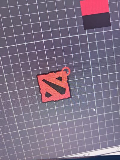 The image shows a digital rendering of the Dota 2 logo, rendered in red with a distressed texture, placed on a grey grid background. A small red loop is attached to the top right corner of the logo. In the upper right corner of the image, a pink rectangle is positioned above a dark grey rectangle. The image does not contain any information about Cheremkhovo, Russia, people, or specific actions.